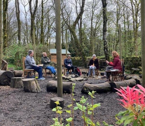 Sarah Hunter teaching a group as part of her nature journalling workshop. They are sat on chairs aaround what looks like an open campfire. 